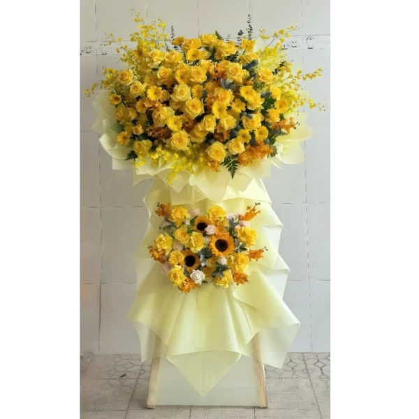 Two tiers of yellow flowers including roses, sunflowers, and orchids, on a light yellow stand.