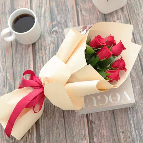 Bouquet of red roses wrapped in cream paper and tied with a red ribbon, next to a mug of coffee.