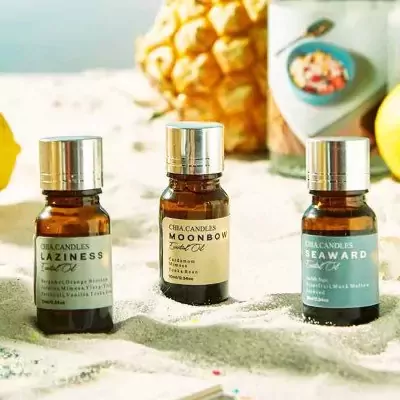 Three bottles of essential oils from C&A Candles with scents "Laziness," "Moonbow," and "Seaward" on sand.