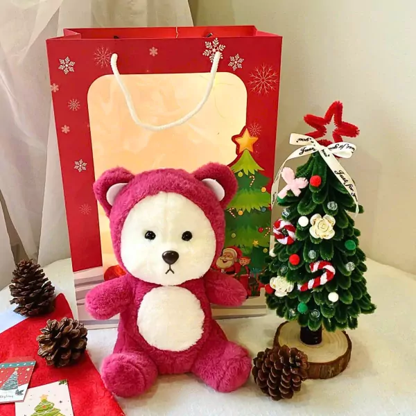 Pink teddy bear with hood, small Christmas tree, pine cones, and red gift bag.
