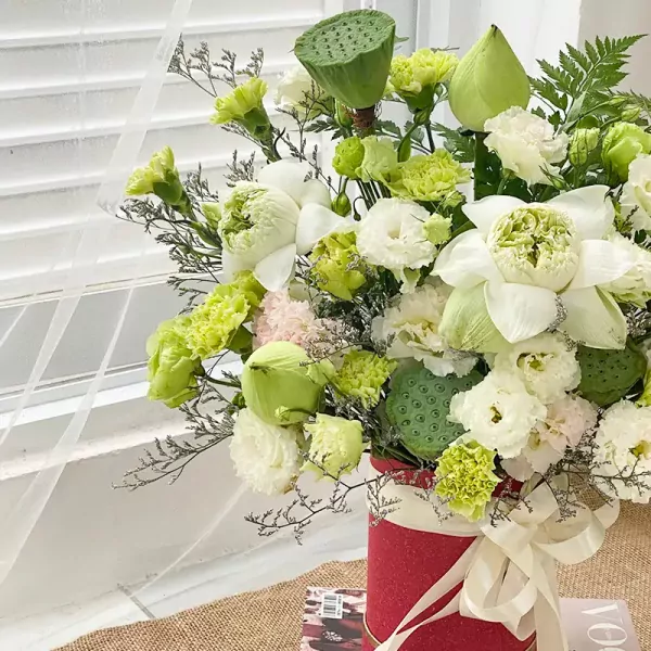 A red box overflowing with white lotuses, green carnations, lisianthus, and lotus seed pods.