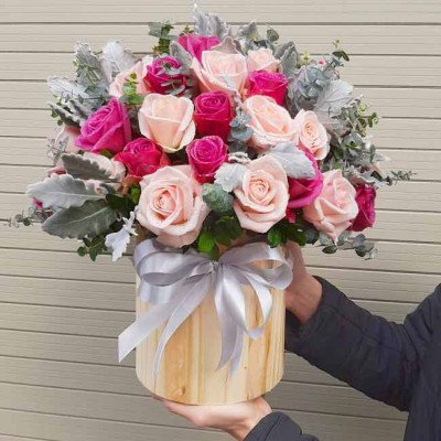A wooden round box filled with pink and magenta roses, accented with dusty miller leaves and a silver ribbon.