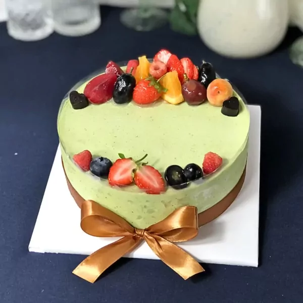Green matcha yogurt mousse cake topped with fresh berries and fruit, tied with a gold ribbon.