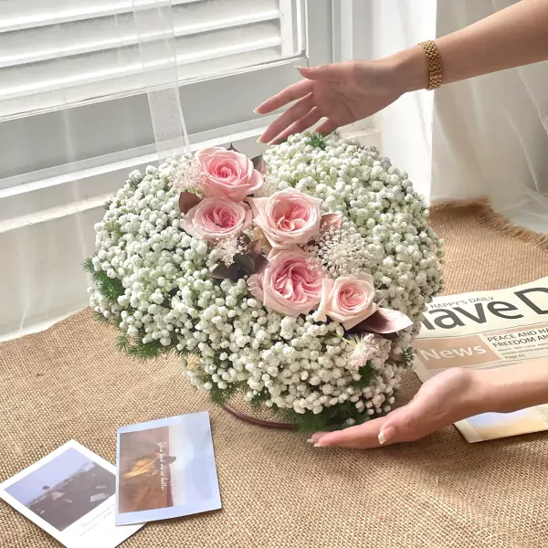 Pink roses and baby's breath arranged in a round bouquet on a burlap cloth, next to photos and a newspaper.