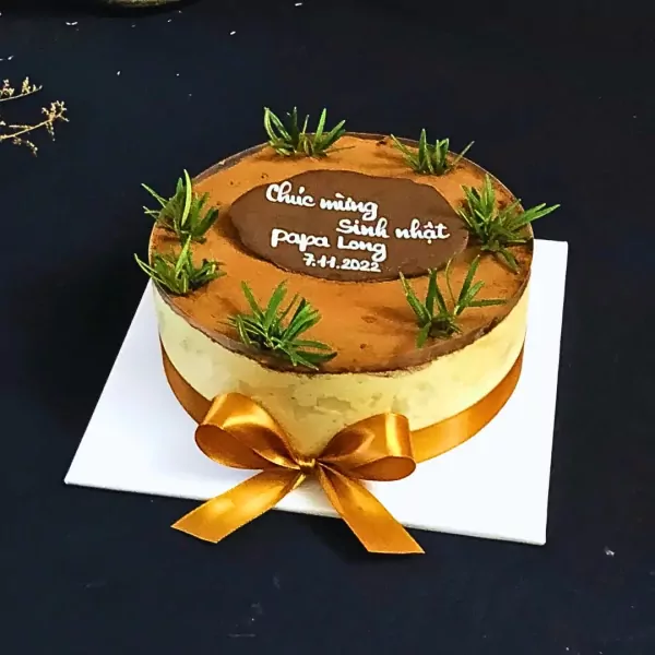 Tiramisu cake decorated with grass, brown frosting, and a gold ribbon.
