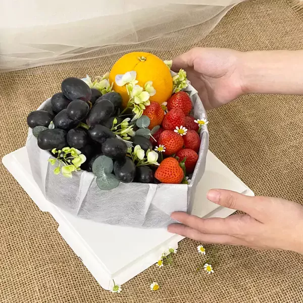 Heart-shaped fruit box with grapes, strawberries, and an orange, decorated with flowers and greenery.