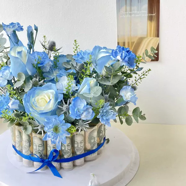 Blue and white floral arrangement in a box decorated with rolled banknotes, tied with a blue ribbon.