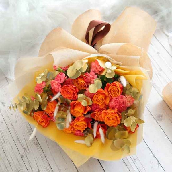 Bouquet of orange roses and pink carnations with eucalyptus, wrapped in light yellow paper.