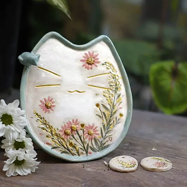 Ceramic cat plate with pink flowers and greenery, next to white daisies.