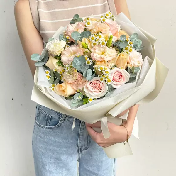 A woman in jeans holds a bouquet of peach and pink roses, lisianthus, and daisies.