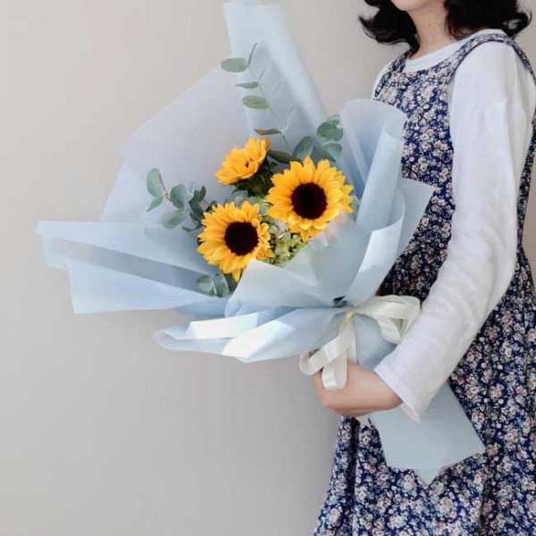 A bouquet of sunflowers and filler flowers wrapped in light blue paper, held by a person in a floral dress.