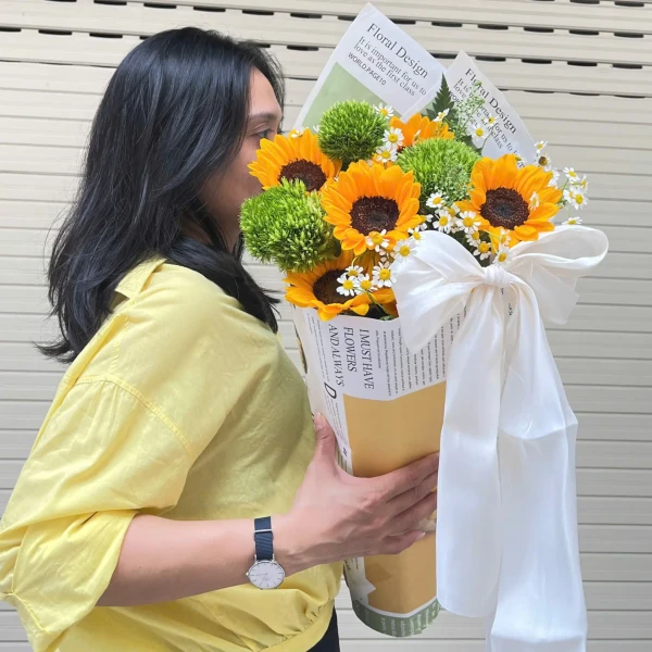 Woman in yellow shirt holding a bouquet of sunflowers, green flowers, and white daisies with a large white ribbon.