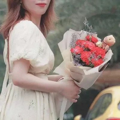 A woman in a cream dress holds a bouquet of red carnations and a mini teddy bear.