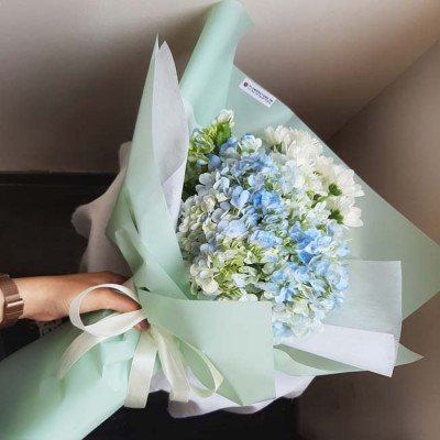 A bouquet of blue hydrangeas and white daisies wrapped in light green and white paper.