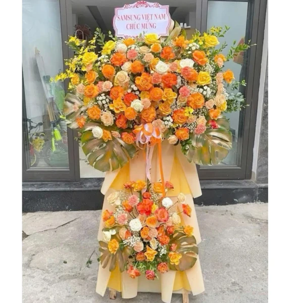 Grand flower stand for grand opening with orange and yellow roses, lilies, and monstera leaves.