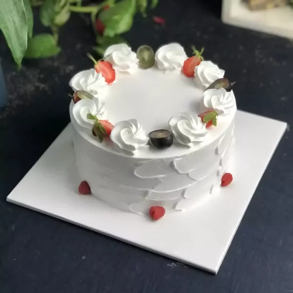 A white fruit cake topped with strawberries, grapes, and whipped cream, on a white cake board.
