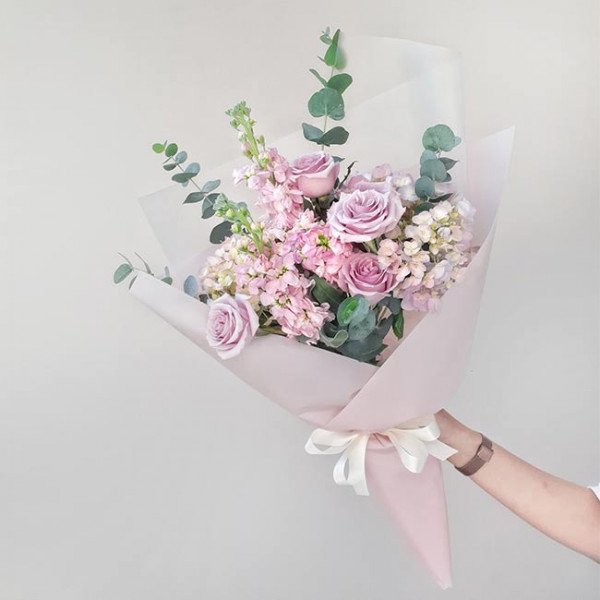 A pastel bouquet of roses, hydrangeas, and snapdragons wrapped in pink paper, held by a person.