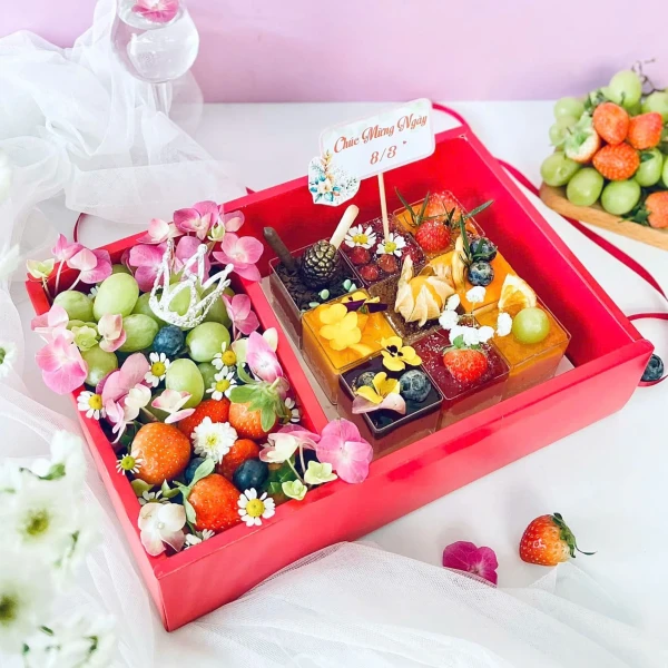 A red gift box filled with small cakes, fresh strawberries, blueberries, and grapes, decorated with flowers.