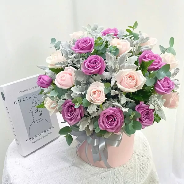 Pink and purple roses in a pink box with a silver ribbon and a book.