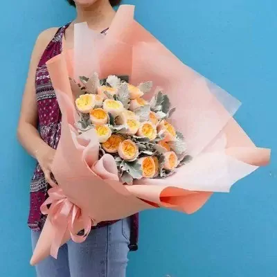 A bouquet of yellow and orange David Austin roses with grey-green foliage wrapped in peach and white paper, tied with a pink ribbon.