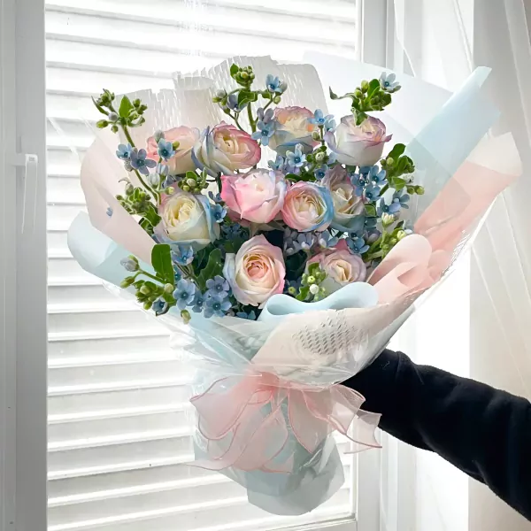 Rainbow roses and blue accent flowers in a bouquet, wrapped in pastel paper and tied with a pink ribbon.