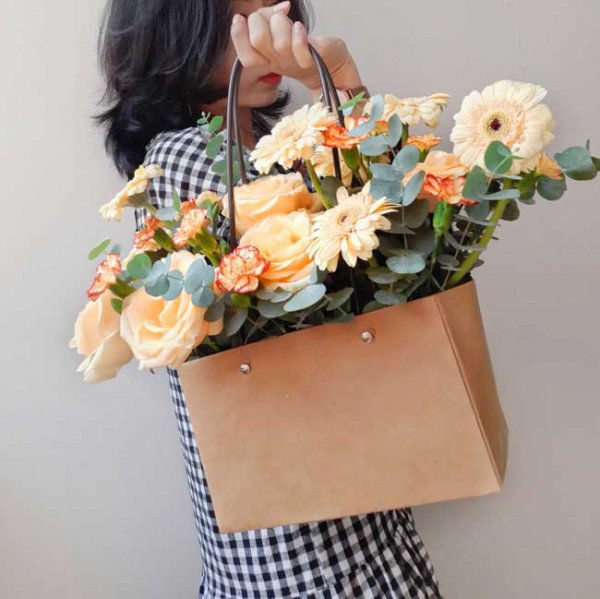 A person holds a brown paper gift bag filled with peach roses, orange gerberas, and carnations.