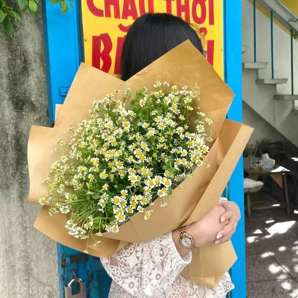 Woman holding a bouquet of small white and yellow daisies wrapped in brown paper.