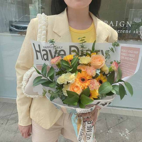 Woman holding a bouquet of yellow and orange flowers wrapped in newspaper and decorated with a heart-shaped bag.