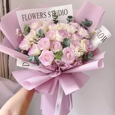 A bouquet of pink and white roses with purple accents, wrapped in lilac paper with a white ribbon.