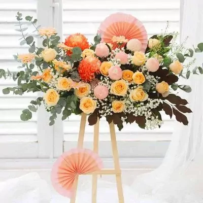 Flower arrangement on easel with orange chrysanthemums, yellow roses, pink ping pong mums, and baby's breath.