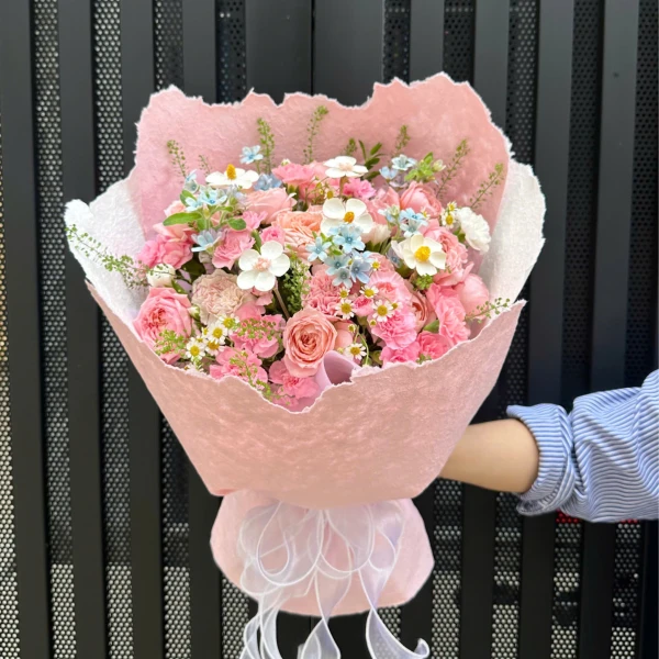 Pink bouquet with roses, carnations, and small blue and white flowers, wrapped in pink paper with a white ribbon.