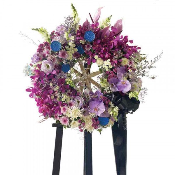 Funeral wreath with purple and white flowers, blue accents, and a black ribbon, displayed on a black easel.