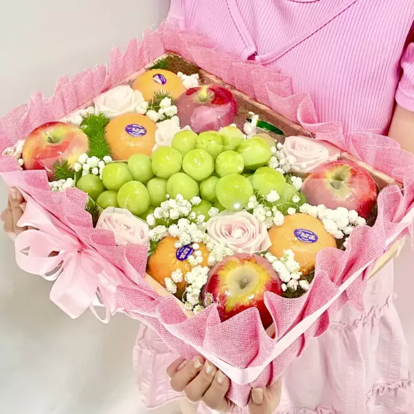 Fruit basket with green grapes, oranges, apples, and pink roses, wrapped in pink mesh.