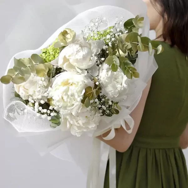 White peonies in a bouquet with eucalyptus and baby's breath, wrapped in white mesh, held by a person in a green dress.