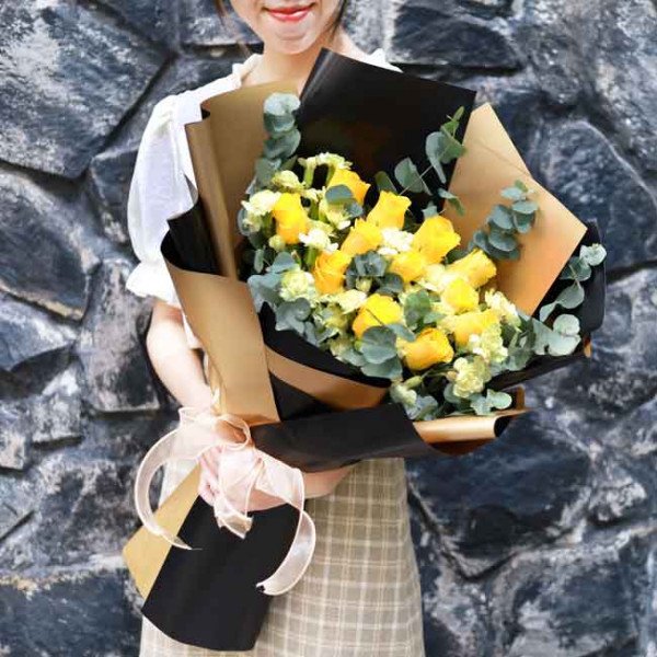 Woman holding a bouquet of yellow roses and baby's breath, wrapped in brown and black paper with a pink bow.