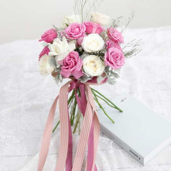 Bridal bouquet of pink and white roses with ribbon streamers on a lace cloth next to a light blue book.