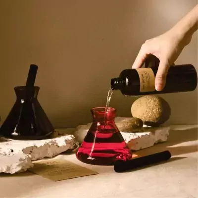 Hand pouring red liquid from amber bottle into red flask, minimal mist diffuser.