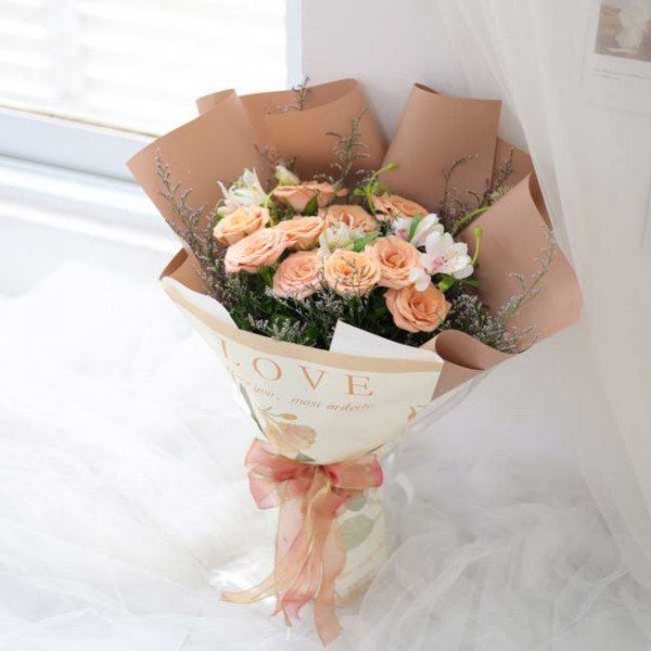 Bouquet of peach roses, white flowers and greenery wrapped in brown and patterned paper with a pink ribbon.