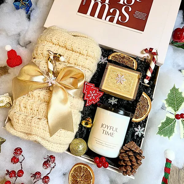 Christmas gift box with knitted scarf, candle, music box, dried oranges, pinecone, and festive ornaments.