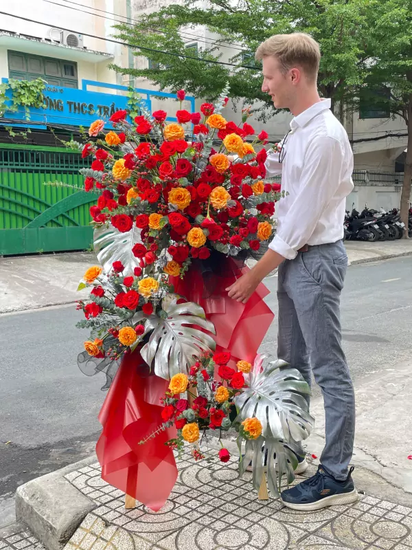 Man in white shirt and grey pants next to a large red and orange rose flower arrangement.