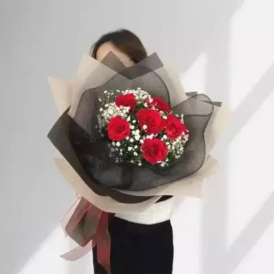 A bouquet of red roses with baby's breath, wrapped in black and beige paper, held by a person.