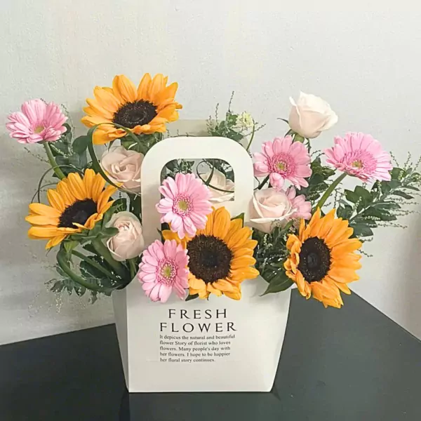 A white floral arrangement with sunflowers, pink gerbera daisies, and light pink roses in a tote bag.