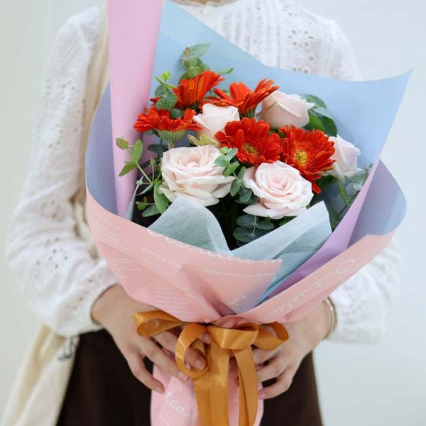 Buket bunga mawar krim dan gerbera oranye cerah dibungkus kertas merah muda dan biru pastel dengan pita emas.