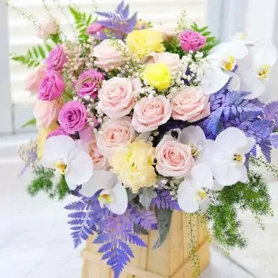 A floral arrangement with pink and yellow roses, white orchids, and purple accents in a wooden box.