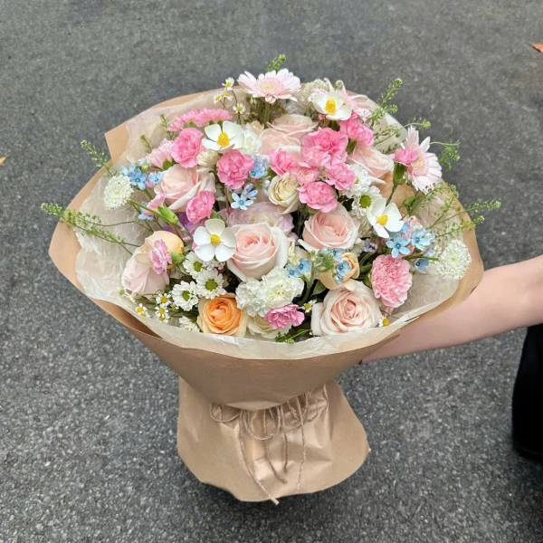 Pink and white roses and carnations with small blue and white flowers in brown paper wrap.