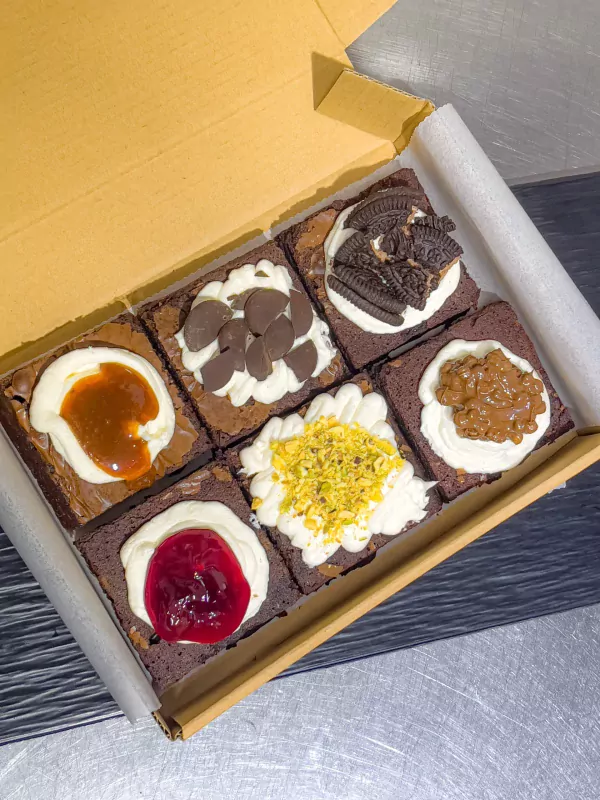 Six gourmet brownies in a box, each topped with different decorations like Oreos, caramel, pistachios, and cherries.