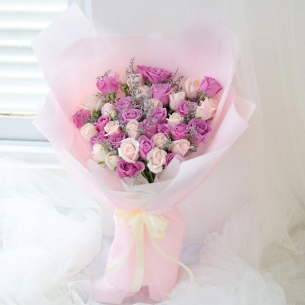 Bouquet of pink and purple roses wrapped in pastel pink paper, tied with a yellow ribbon.