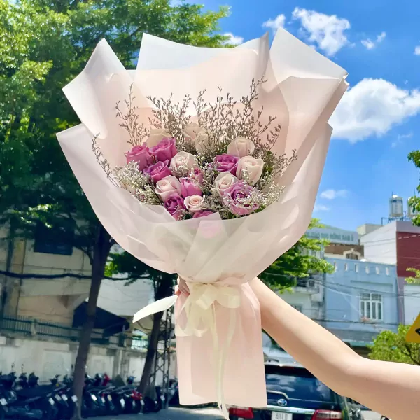 A hand holds a bouquet of pink and white roses, wrapped in delicate pink paper with a ribbon.