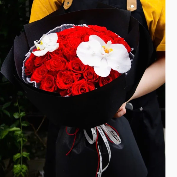 A bouquet of red roses with two white orchids, wrapped in black paper and tied with a white ribbon.