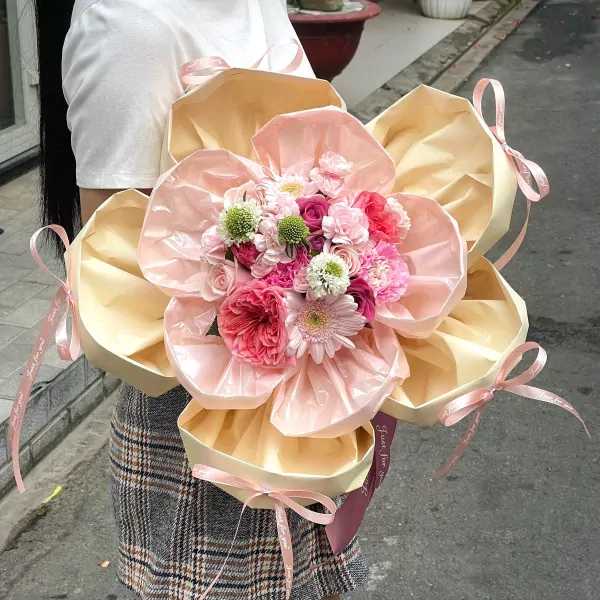 A bouquet of pink and cream flowers in a layered light pink and cream wrapper, tied with ribbons.